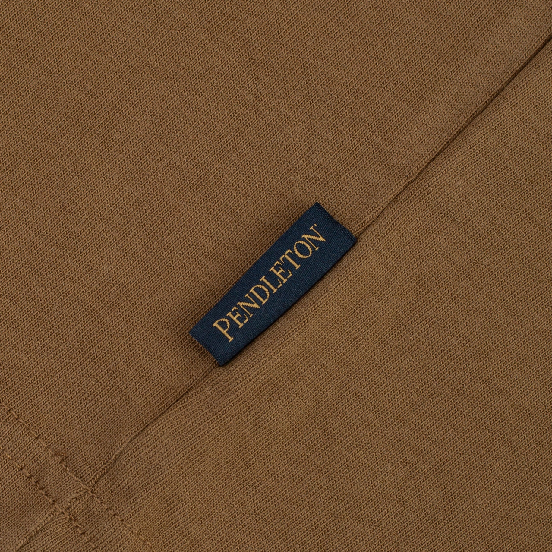 Close-up of a brown woven fabric with a small dark navy label stitched on it, printed with the brand name PENDLETON in gold lettering.